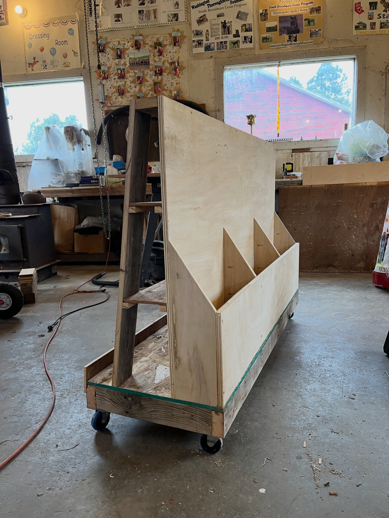 A wooden storage unit with shelves is positioned on casters in a workshop setting.