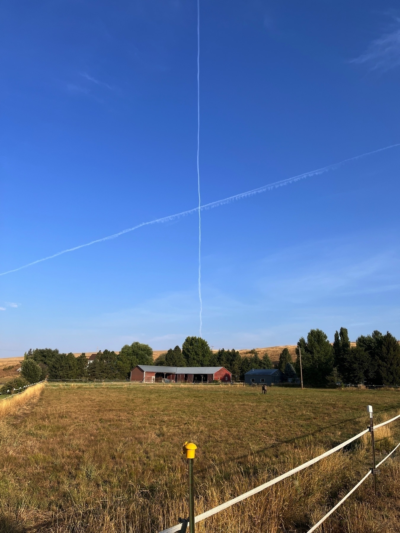 A contrail forms an X shape in a clear blue sky over a rural landscape with a house and open field.