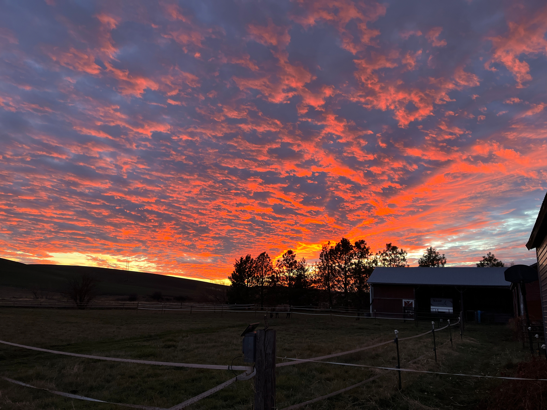 A vibrant sunset fills the sky with hues of orange and red above a rural landscape featuring a barn and trees.